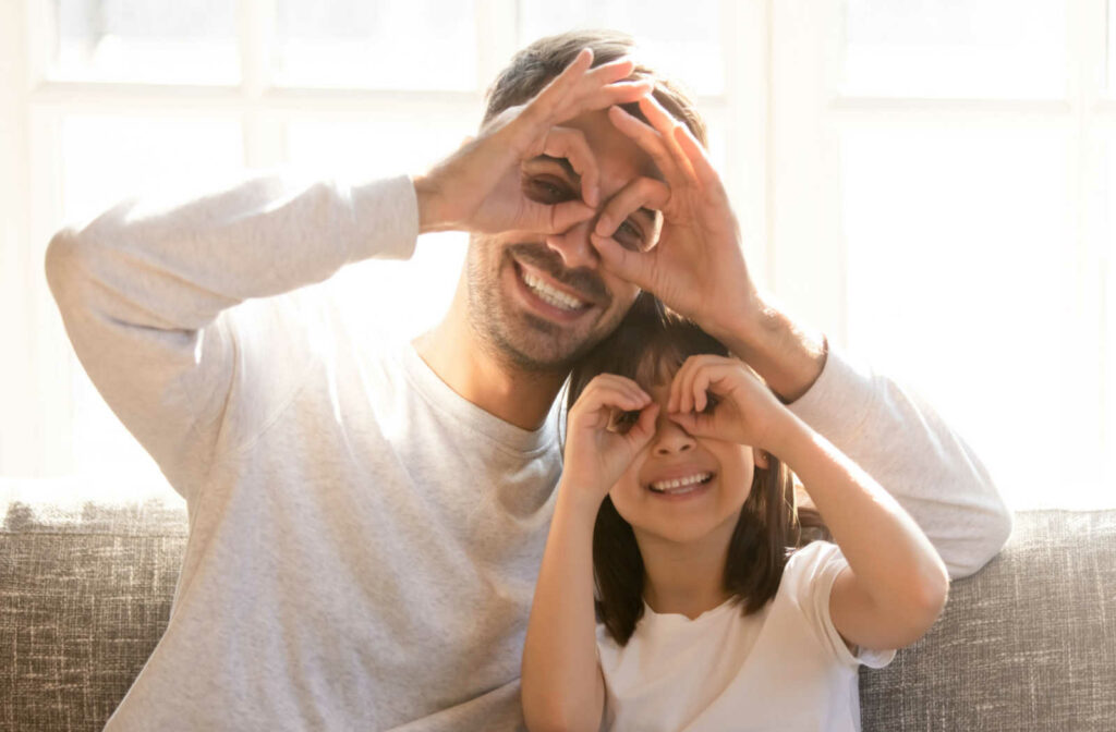 A father and daughter sitting on a couch smiling while looking through circles made with their fingers held up their eyes.