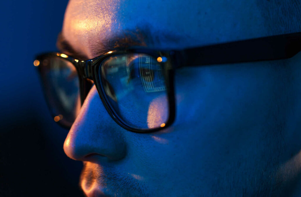 A close up of a younger male wearing blue light glasses showing a reflection of a computer screen in the lens.