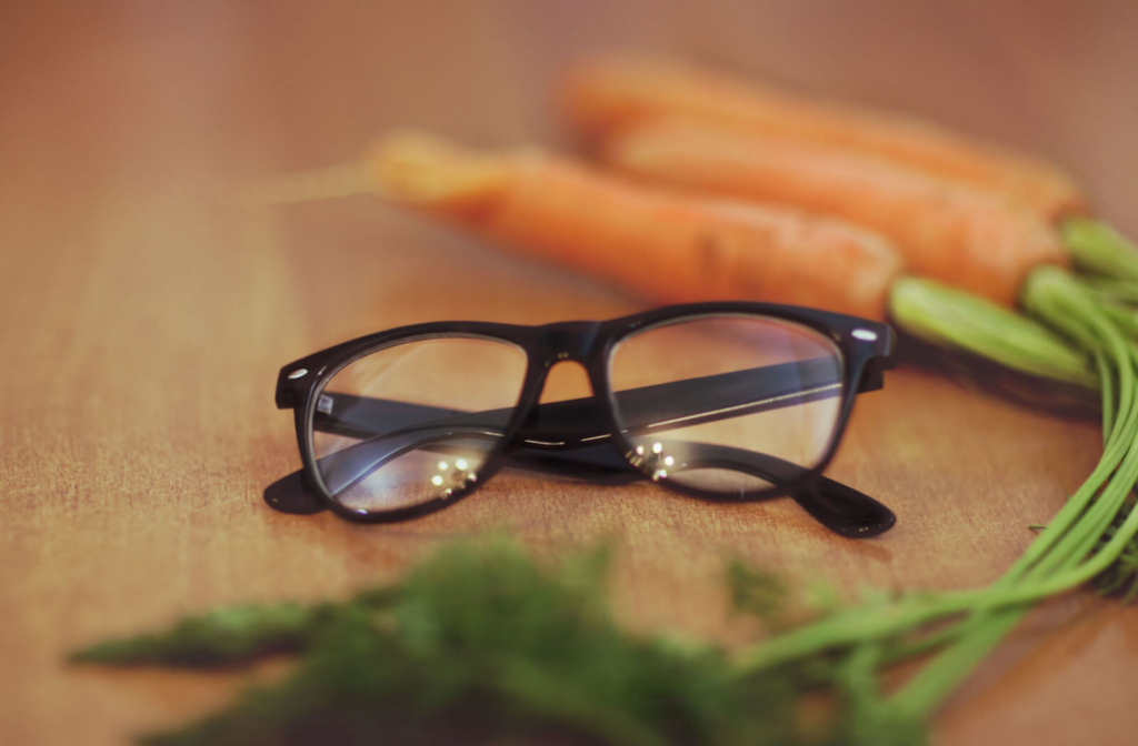 A pair of eyeglasses resting next to fresh carrots on a wooden surface.