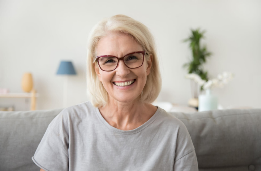 A senior woman with eyeglasses sitting on a couch smiling, looking directly at the camera.