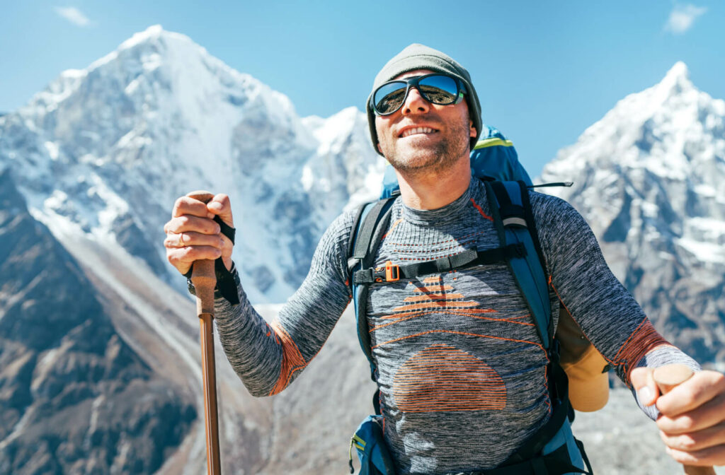A man on top of a mountain wearing UV protection sunglasses and a backpack while hiking with poles.