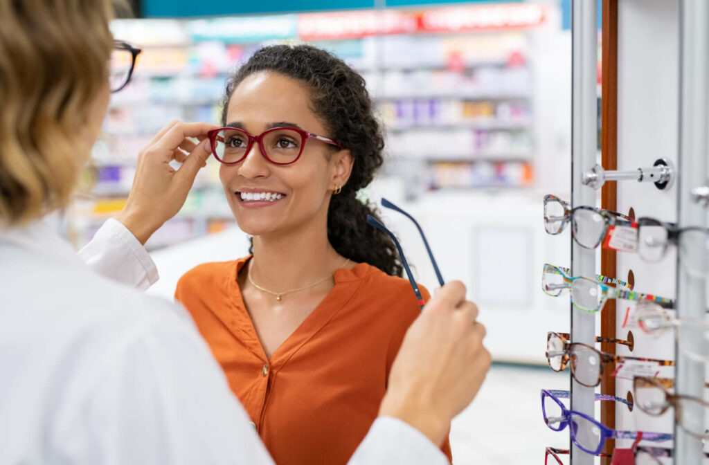 A young woman smiling and trying on glasses in a store while being assisted by an optician or optometrist.