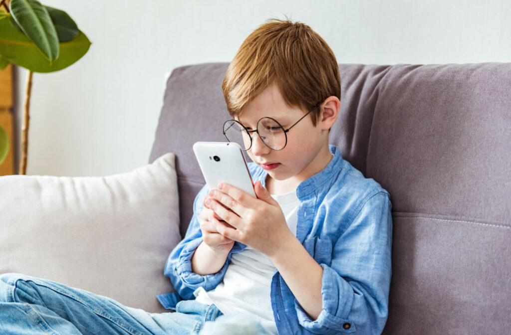 A child with glasses, sitting on a couch and holding his smartphone very close to his face.
