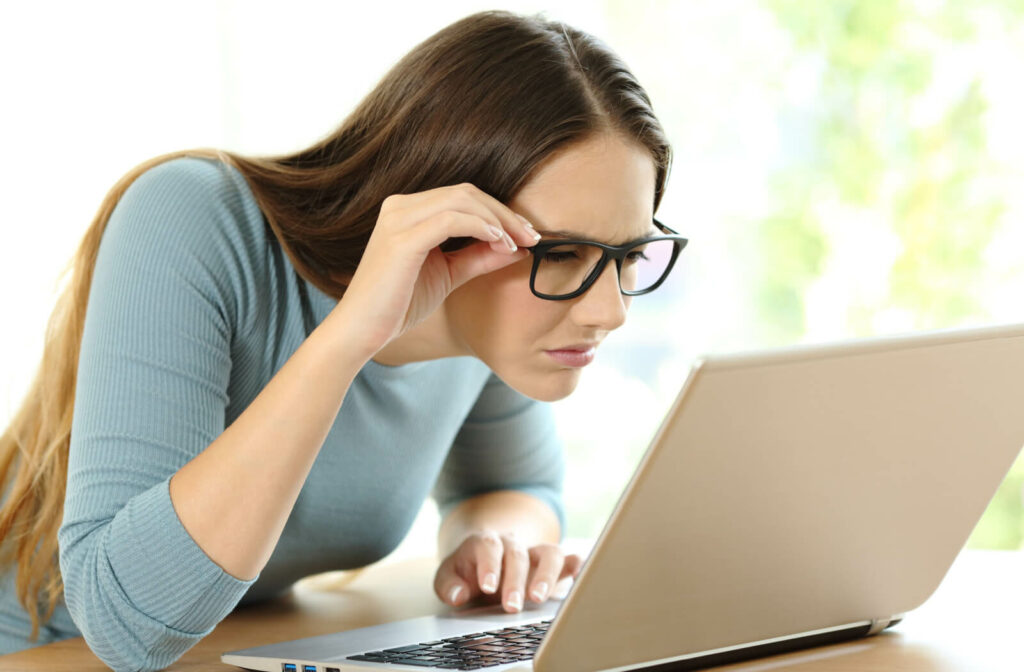 A woman sitting in front of a laptop and adjusting her glasses as she squints to see.