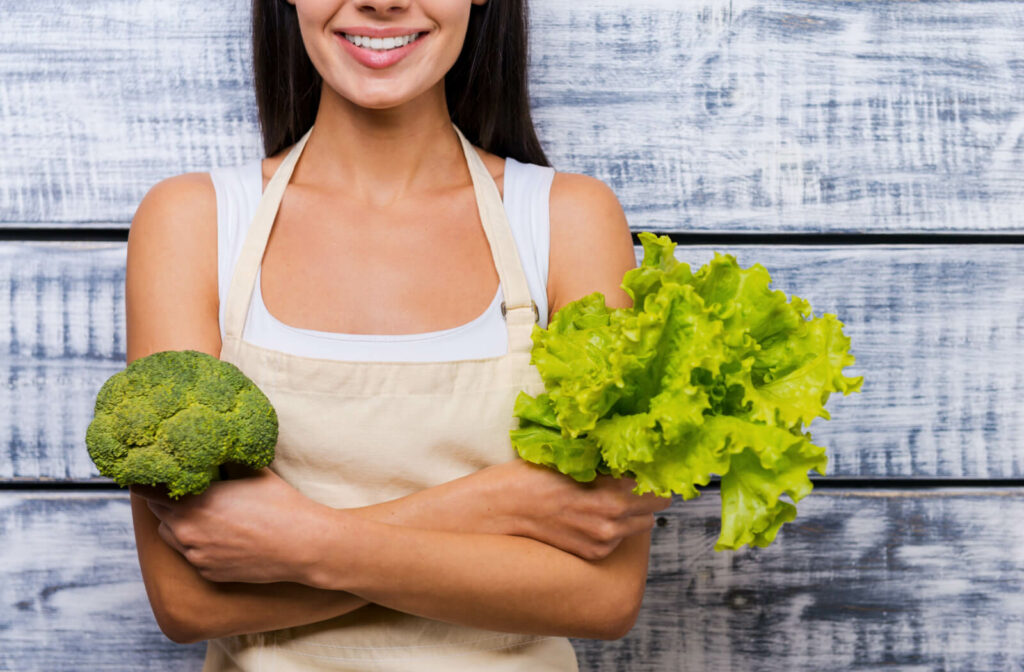A woman smiling woman with apron and holding a lettuce and broccoli while standing in front of wooden background.