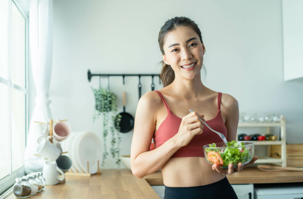 A fit young woman holding a bowl of salad while smiling and looking directly at the camera.