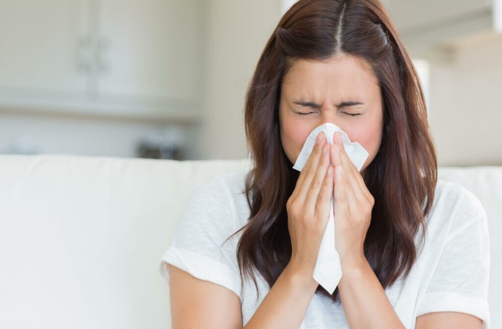 A woman seated on a couch in the living room, sneezes while holding a tissue.
