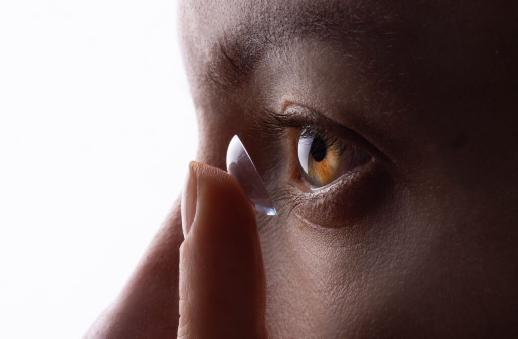 Close-up of a person's profile with a hazel eye, holding a contact lens on their fingertip near the eye.